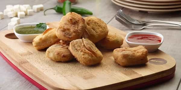 Paneer pakora on a wooden board with green chutney and tomato sauce, garnished with spices.