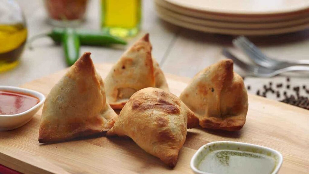 Four samosas on a wooden board with dipping sauces, ready to eat. Samosa (4 pcs).